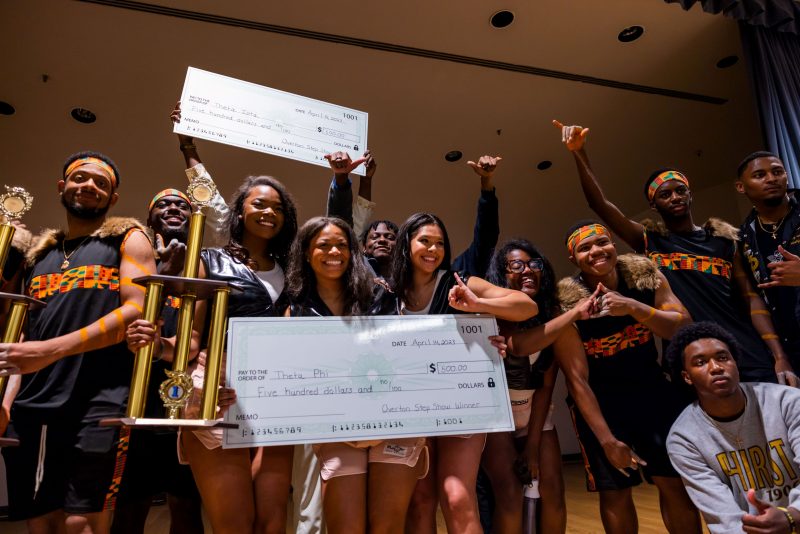 Several students in their performance costumes hold up trophies and large checks that they received as prizes. 
